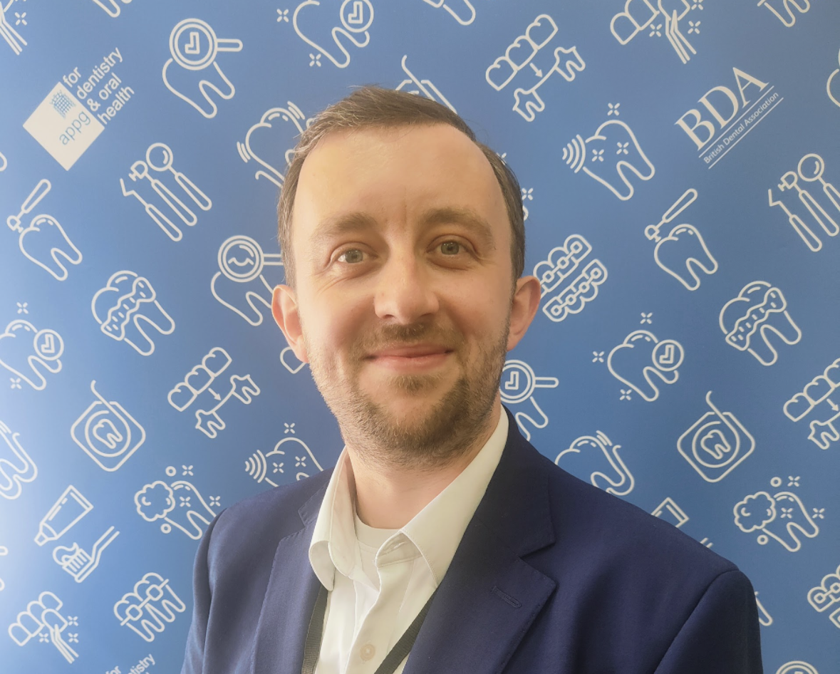A smiling man in a suit stands in front of a blue background with dental icons.